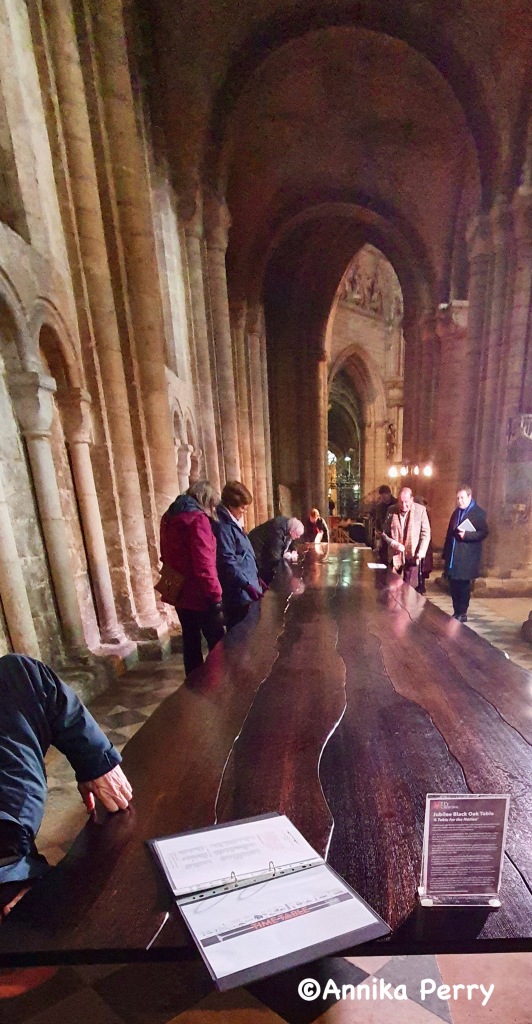 "My photo of the Jubilee Oak Table in Ely Cathedral. It is taken from one end and one can see the full length down. At the furthest end a group of visitors are gathered, touching the table, peering underneath, reading information leaflets. The table is set in the expanse of the cathedral with lofty stone arches all around."