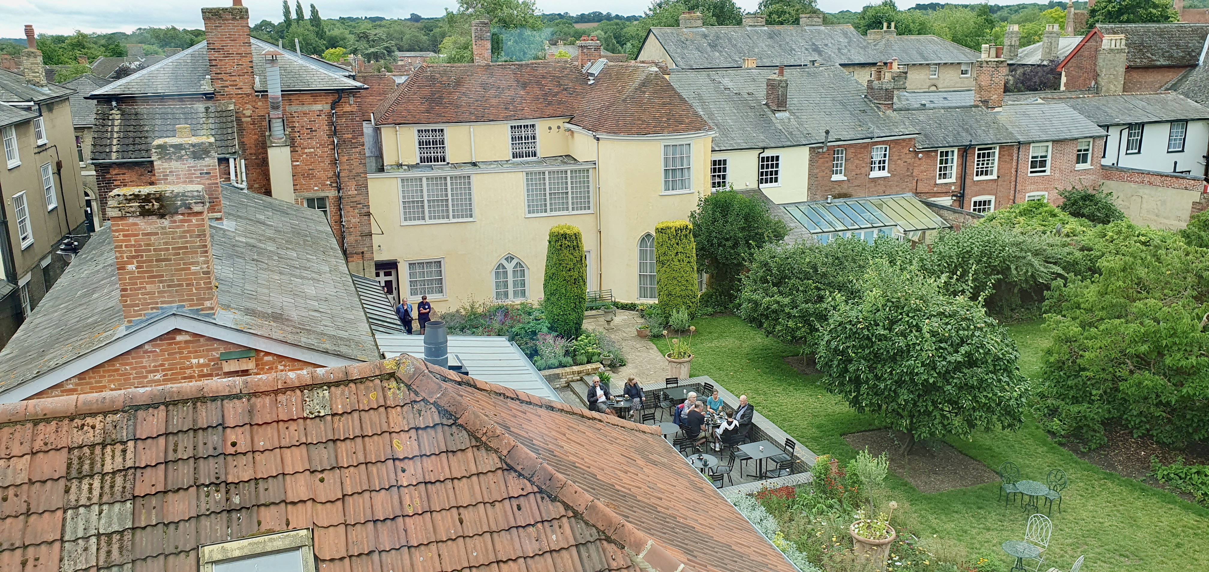 "View looking down onto the rear yellow of Gainsborough house, set behind pretty garden with patio seating area for cafe to the left."