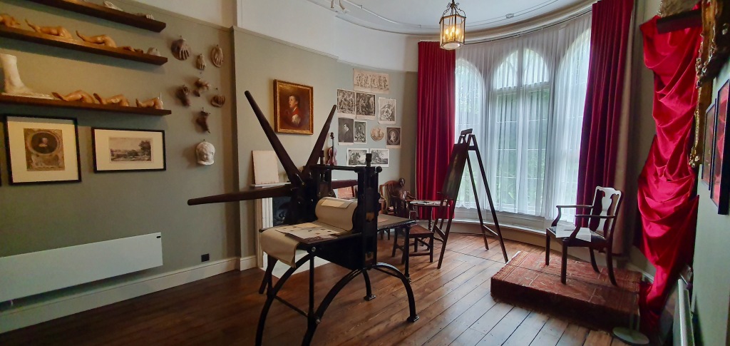 "Shelving to the right with plaster casts of hands and feet, in the centre of the painting room the press and easel and to the right sitters chair with red silk backdrop."