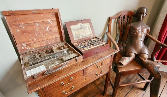 "Wooden wooden chest of drawers and on top two boxes, one of traditional  painting tools the other of painting blocks. A metal manaqun rest on a chair next to this."