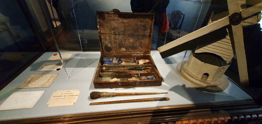 "Constable's old paint blox and brushes next to model of one of many wooden windmills from the area."