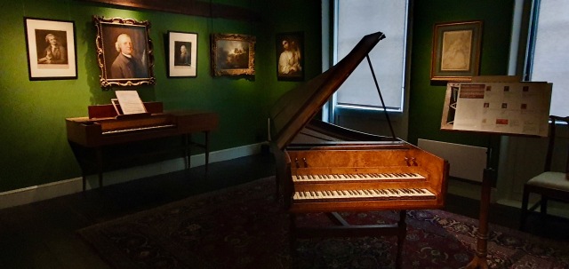 "Harpsicord, like a grand piano but smaller, the top lid open and on the green walls many paintings."